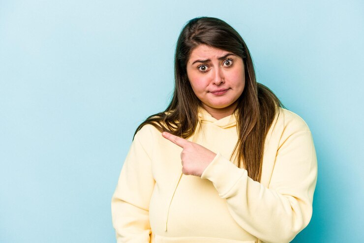 फोटो young caucasian overweight woman isolated on blue background smiling and pointing aside showing something at blank space