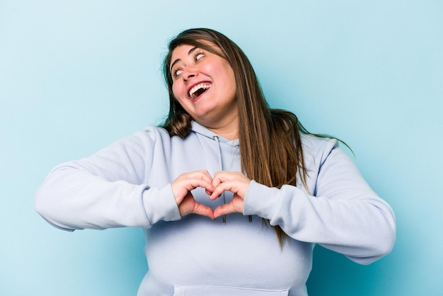 Young caucasian overweight woman isolated on blue background smiling and showing a heart shape with hands