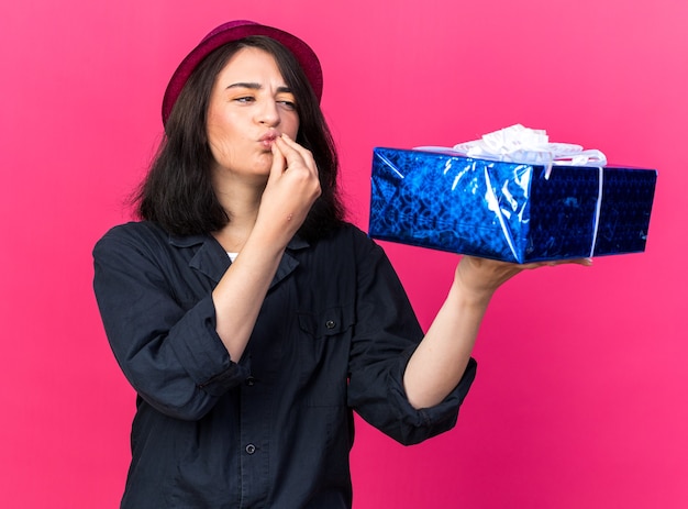 Young caucasian party girl wearing party hat holding and looking at gift package doing chef's kiss gesture isolated on pink wall