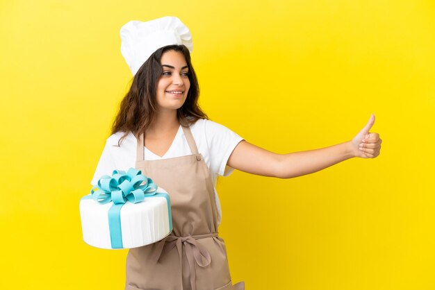 Young caucasian pastry chef woman with a big cake isolated on yellow background giving a thumbs up gesture