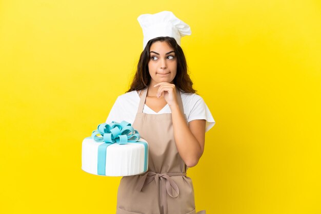 Young caucasian pastry chef woman with a big cake isolated on yellow background having doubts and thinking