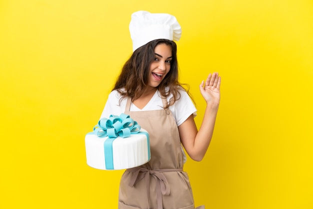 Young caucasian pastry chef woman with a big cake isolated on yellow background saluting with hand with happy expression