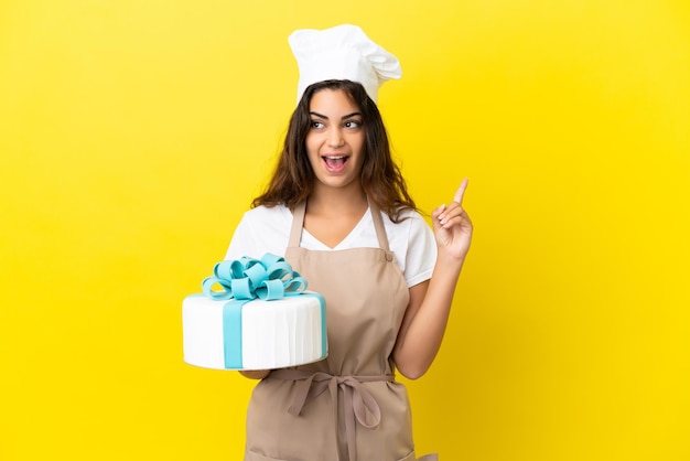 Young caucasian pastry chef woman with a big cake isolated on yellow background thinking an idea pointing the finger up