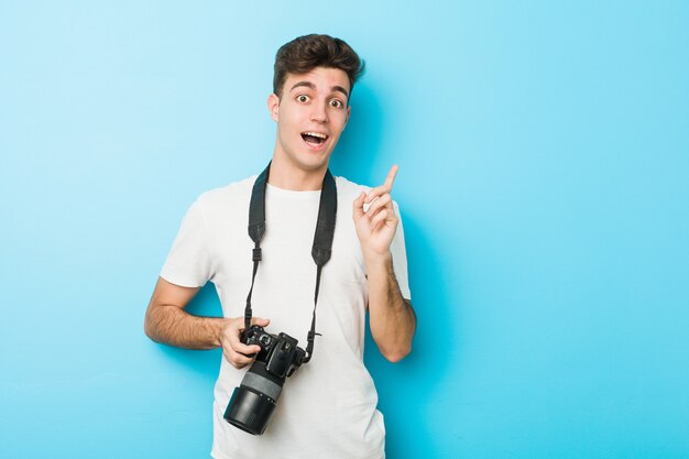 Young caucasian photographer man holding a camera smiling cheerfully pointing with forefinger away.