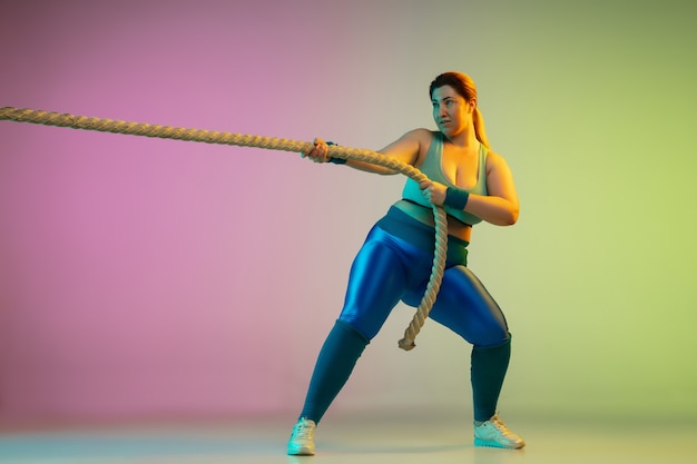 Young caucasian plus size female model's training on gradient purple green wall in neon light. Doing workout exercises with ropes. Concept of sport, healthy lifestyle, body positive, equality.