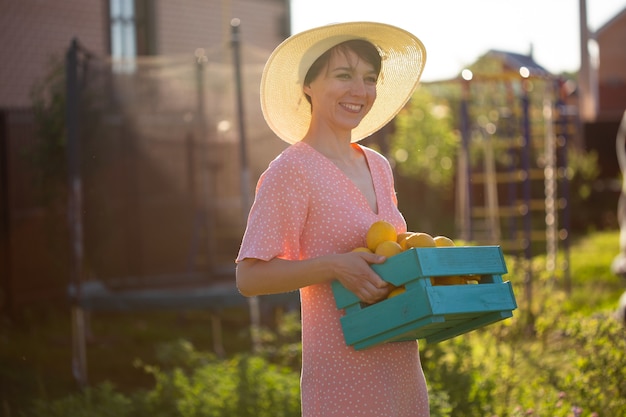 Il giovane giardiniere caucasico della donna positiva in cappello e vestito dentellare tiene la scatola con i limoni nelle sue mani il giorno di estate pieno di sole. concetto di agricoltura e giardinaggio biologico