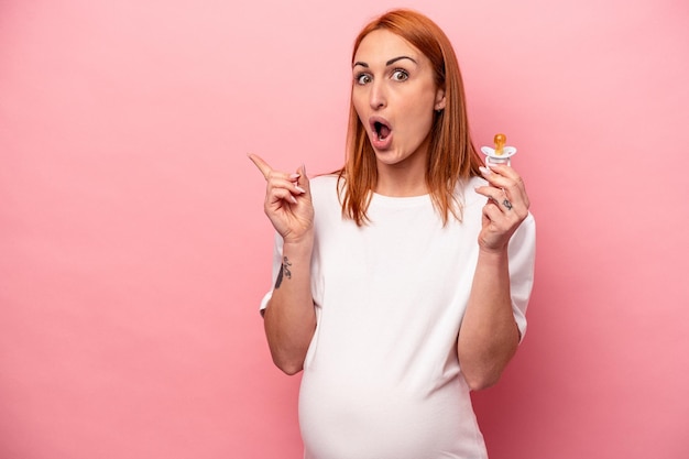 Young caucasian pregnant woman holding pacifier isolated on pink background pointing to the side