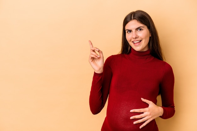 Молодая кавказская беременная женщина, изолированных на бежевом фоне, весело улыбаясь, указывая указательным пальцем.