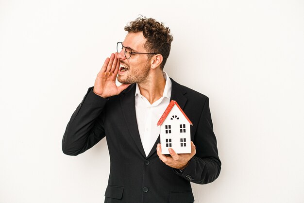 Young caucasian real estate holding a toy home isolated on white background shouting and holding palm near opened mouth.