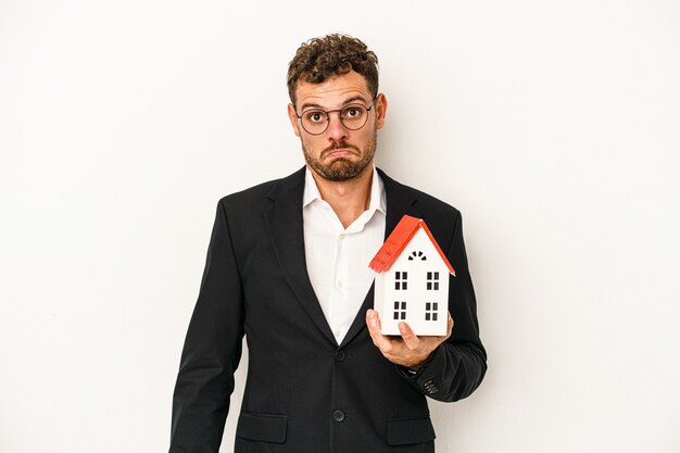 Young caucasian real estate holding a toy home isolated on white background shrugs shoulders and open eyes confused.