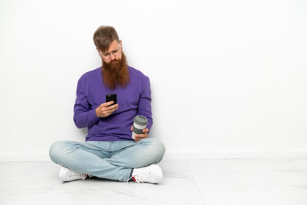 Young caucasian reddish man sitting on the floor isolated on white background holding coffee to take away and a mobile