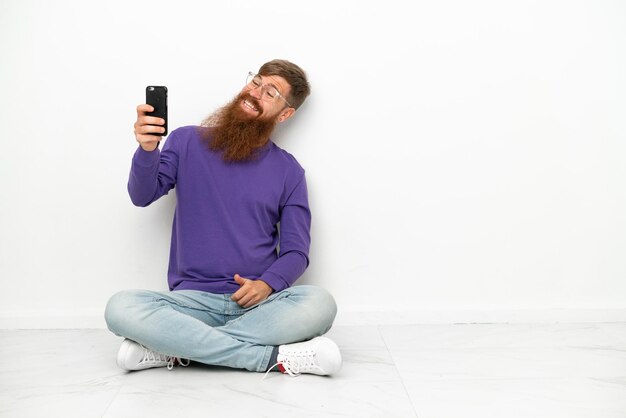 Young caucasian reddish man sitting on the floor isolated on white background making a selfie