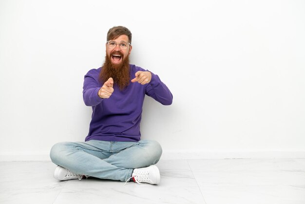 Young caucasian reddish man sitting on the floor isolated on white background pointing to the front and smiling