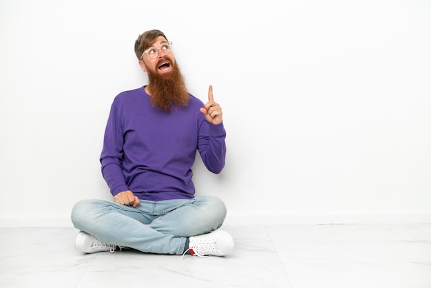 Young caucasian reddish man sitting on the floor isolated on white background pointing up and surprised