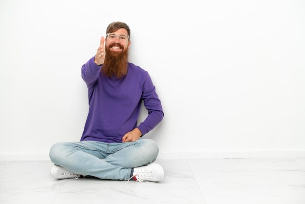 Young caucasian reddish man sitting on the floor isolated on white background shaking hands for closing a good deal