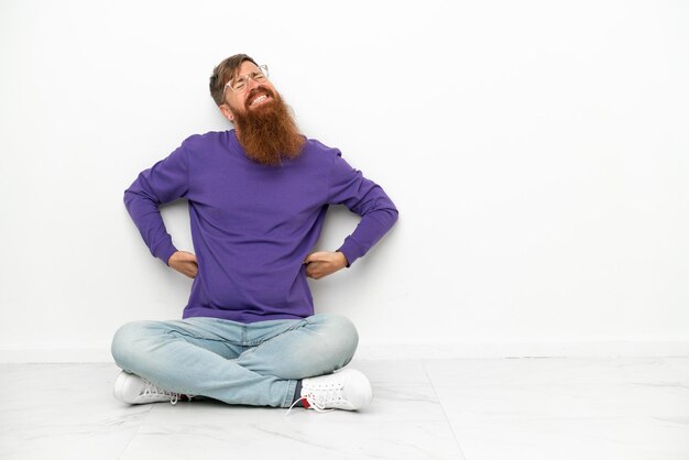 Young caucasian reddish man sitting on the floor isolated on white background suffering from backache for having made an effort