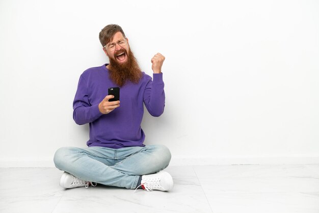 Young caucasian reddish man sitting on the floor isolated on white background with phone in victory position