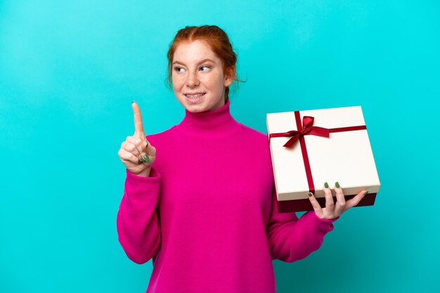 Foto giovane donna rossastra caucasica che tiene un regalo isolato su sfondo blu che intende realizzare la soluzione mentre solleva un dito