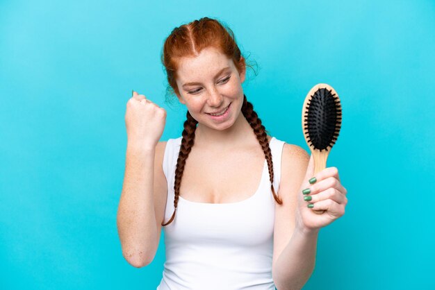 写真 勝利を祝う青の背景に分離された髪の櫛を持つ若い白人赤みを帯びた女性