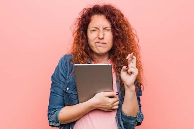 Young caucasian redhead woman holding a tablet crossing fingers for having luck