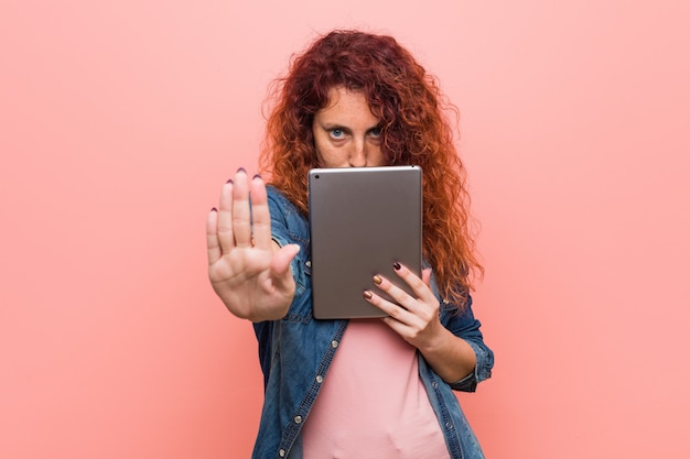 Young caucasian redhead woman holding a tablet standing with outstretched hand showing stop sign, preventing you.