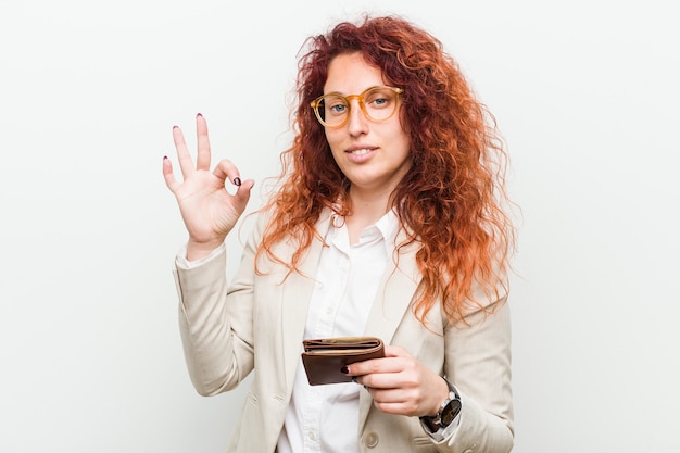 Young caucasian redhead woman holding a wallet cheerful and confident showing ok gesture.