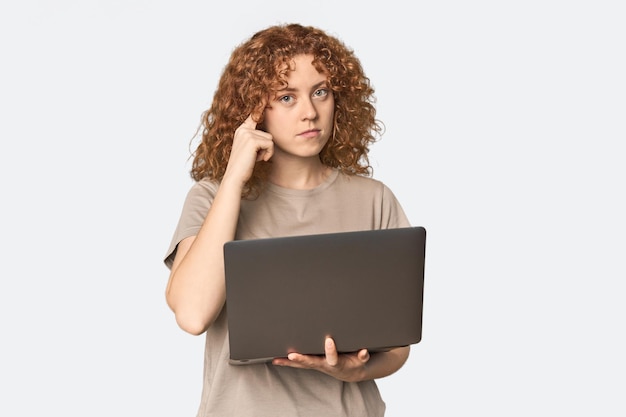 Фото Молодая белокожая рыжая женщина с ноутбуком, указывающим на храм, с пальцем, сосредоточенным на задаче.