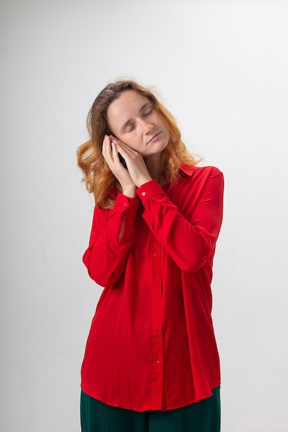 Young caucasian redheaded woman with long hair in red shirt holding hands near her face, pretending to sleep. sleepy female with closed eyes standing on white background