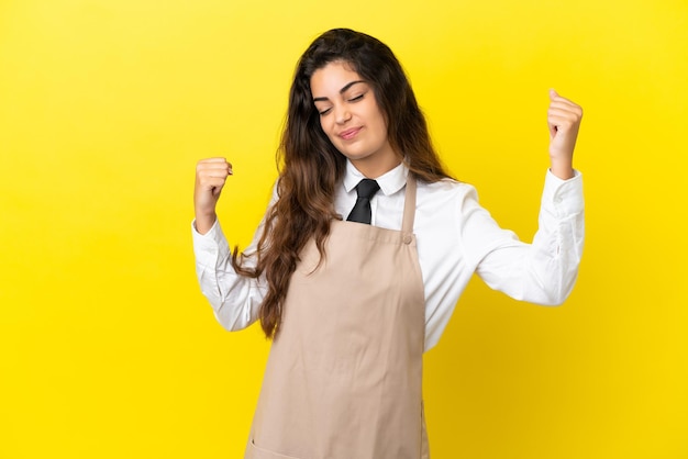 Young caucasian restaurant waiter isolated on yellow background celebrating a victory