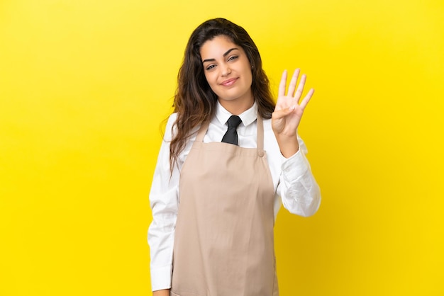 Young caucasian restaurant waiter isolated on yellow background happy and counting four with fingers