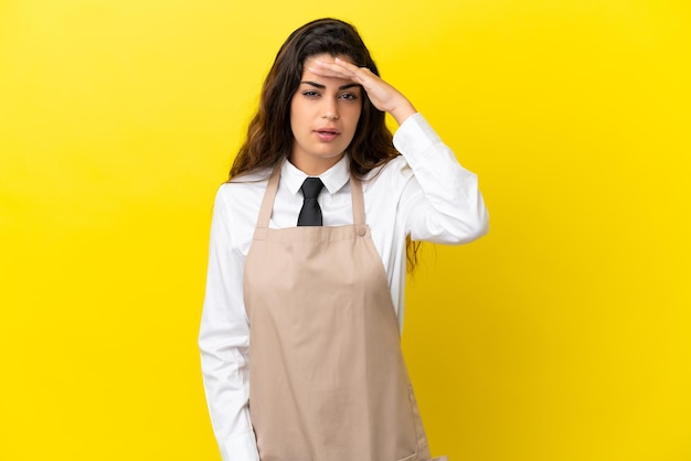 Young caucasian restaurant waiter isolated on yellow background looking far away with hand to look something
