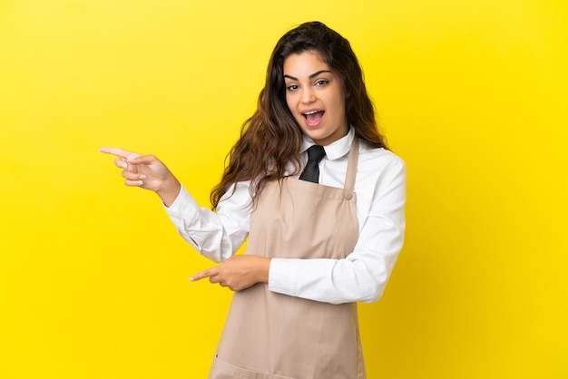 Young caucasian restaurant waiter isolated on yellow background surprised and pointing side