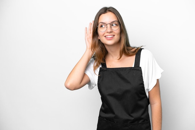 Young caucasian restaurant waiter woman isolated on white background listening to something by putting hand on the ear