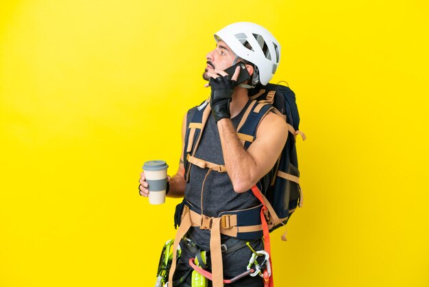 Young caucasian rock climber man isolated on yellow background holding coffee to take away and a mobile