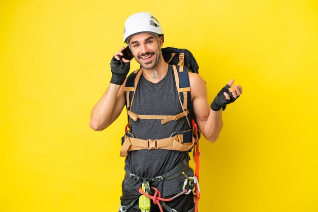 Young caucasian rock climber man isolated on yellow background
keeping a conversation with the mobile phone with someone