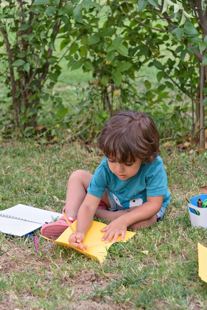 Young caucasian school boy sitting in park outdoor doing school\
homework child kid writing in notebook with pencil outside self\
education learning studying early development for children