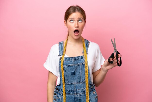 Young caucasian seamstress woman isolated on pink background looking up and with surprised expression