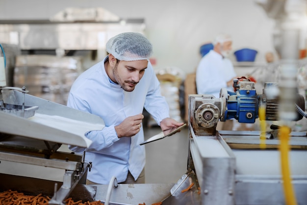 Young Caucasian serious supervisor evaluating quality of food in food plant while holding tablet. Man is dressed in white uniform and having hair net.