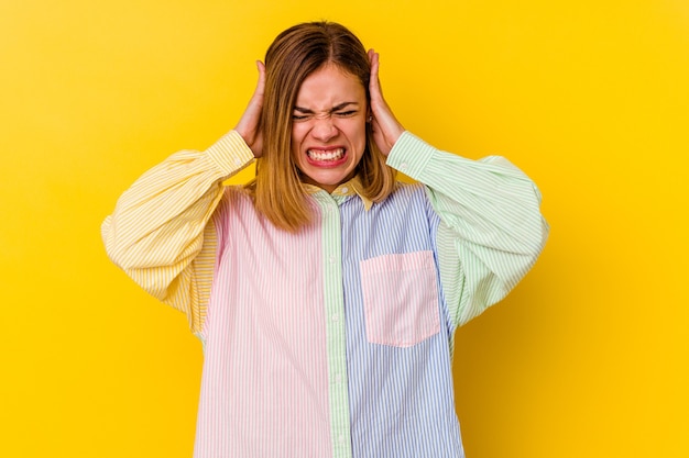 黄色の背景に孤立した若い白人の細い女性は、あまりにも大きな音を聞かないように手で耳を覆っています。