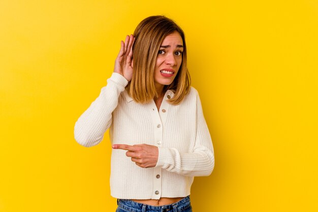 Young caucasian skinny woman isolated on yellow background trying to listening a gossip.