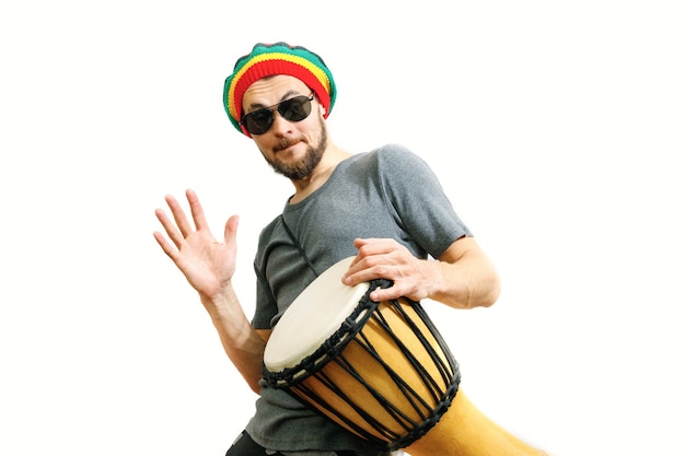 Young caucasian smiling man in rasta hat and sunglasses on white background play on djembe