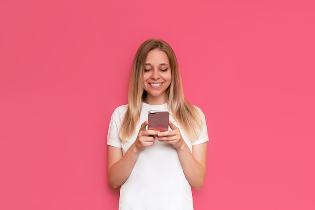 若い白人の笑顔のかわいい金髪の女性が、明るい色のピンクの壁に隔離された画面を見ながら携帯電話を持っている白いTシャツを着た女の子がスマートフォンを使う