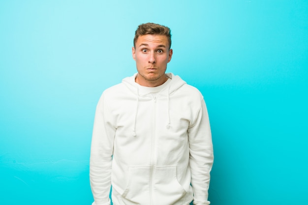 young caucasian sport man shrugs shoulders and open eyes confused.