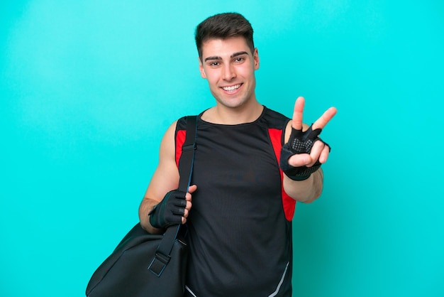 Young caucasian sport man with sport bag isolated on blue background smiling and showing victory sign