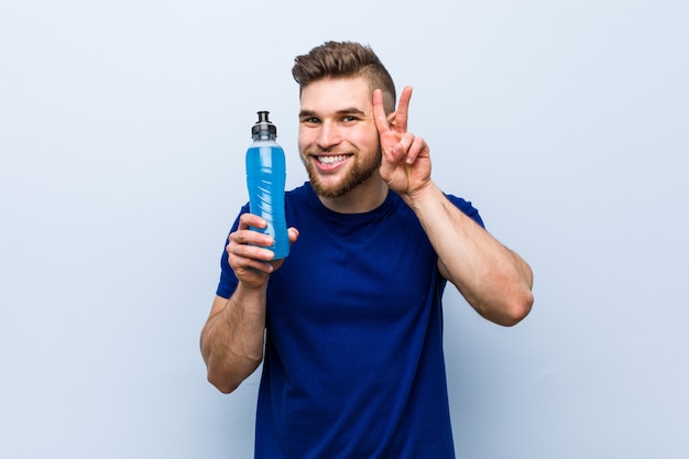 Young caucasian sportsman holding an isotonic drink showing victory sign and smiling broadly.