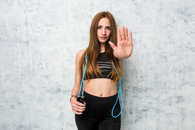 Young caucasian sportswoman holding a jump rope standing with outstretched hand showing stop sign, preventing you.