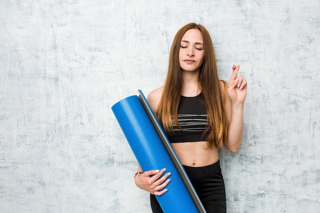 Photo young caucasian sportswoman holding a mat crossing fingers for having luck