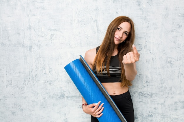 Young caucasian sportswoman holding a mat pointing with finger at you as if inviting come closer.