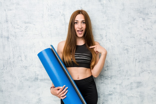 Young caucasian sportswoman holding a mat surprised pointing at himself, smiling broadly.