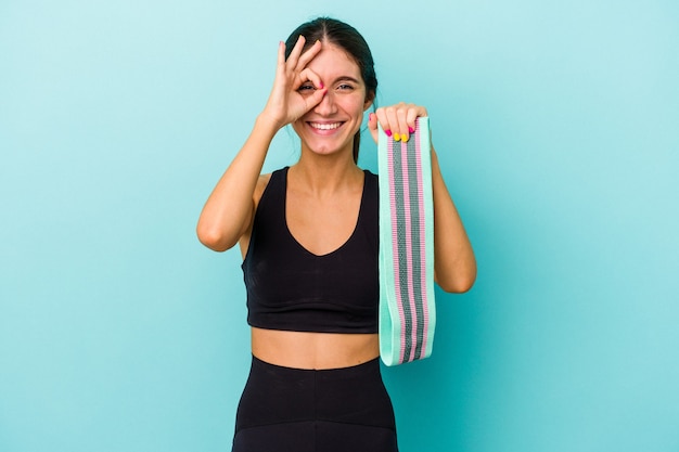 Giovane donna sportiva caucasica che tiene elastici isolati su sfondo blu eccitato mantenendo il gesto ok sull'occhio.